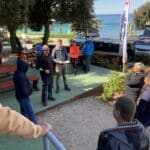 Skupina lidí, někteří stojící a jiní sedící, se shromáždili venku u moře ve Vrsaru v Chorvatsku na setkání nebo akci v roce 2024, s auty a stromy