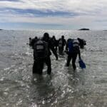 Potápěči vstupující do oceánu s vybavením ve Vrsaru, brodící se mělkou vodou pod jasnou oblohou.