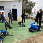 Potápěči v neoprenech připravujících výstroj před potápěčským obchodem ve Vrsaru s potápěčským vybavením rozloženým na zemi.