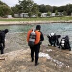 Tři potápěči, kteří se za zataženého dne chystají vstoupit do čistého jezera ve Vrsaru s chatkami v pozadí.