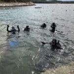 Pět potápěčů ve výstroji, někteří mávají, plují v čisté, mělké vodě poblíž skalnatého pobřeží v Chorvatsku 2024 se žlutými bójemi v pozadí.