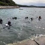 Skupina potápěčů v černých neoprenech se připravuje na potápění v čistém moři poblíž skalnatého pobřeží Chorvatského Vrsaru pod zataženou oblohou.