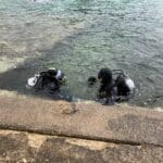 Dva potápěči připravující vybavení v mělké vodě poblíž betonové římsy ve Vrsaru.