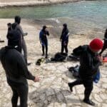 Potápěči v neoprenech připravujících výstroj na skalnatém pobřeží ve Vrsaru, někteří potápěči vstupují do vody a jedna osoba pořizuje fotografie.