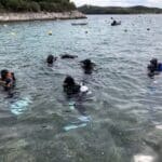 Potápěči ve výstroji vstupují do vody ze skalnatého pobřeží poblíž Vrsaru s několika čluny plujícími poblíž pod zataženou oblohou.