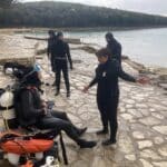 Potápěči v neoprenech s výstrojí na skalnatém pobřeží ve Vrsaru, jeden potápěč sedí s vybavením, další stojí a konverzují u moře.