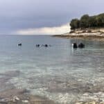Potápěči se připravují na potápění v čistých mělkých vodách poblíž skalnatého pobřeží ve Vrsaru pod zataženou oblohou.