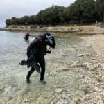 Potápěč nastavuje vybavení při vstupu do vody na skalnaté pláži ve Vrsaru s dalším potápěčem vpředu a stromy v pozadí.