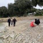 Dva policisté jdou po oblázkové pláži v Chorvatském Vrsaru, zatímco dva lidé se krčí u vody a zkoumají břeh v zalesněné pobřežní oblasti.